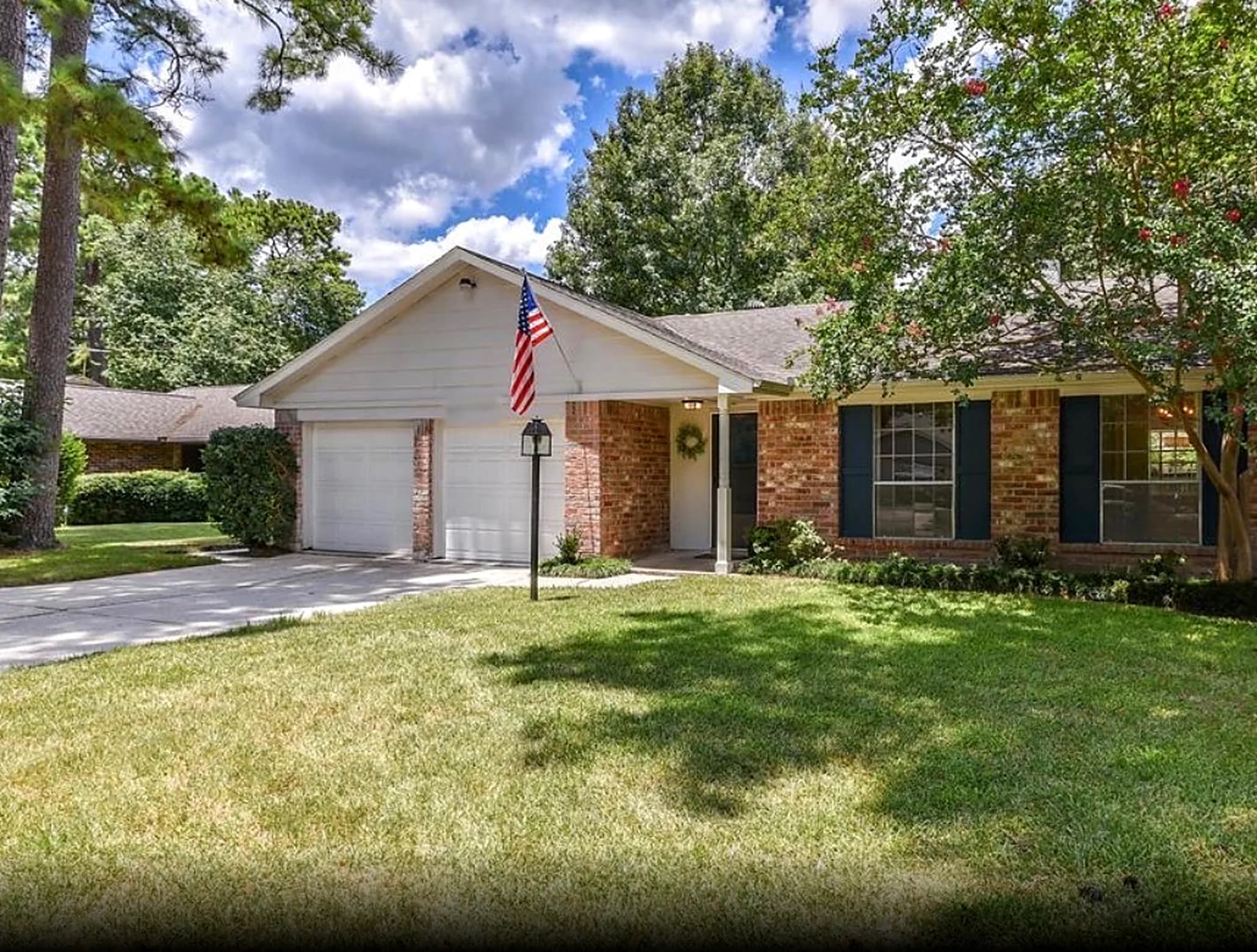 Front of a single story brick house with a two car garage, a flag by the front door and a mowed lawn under tall trees